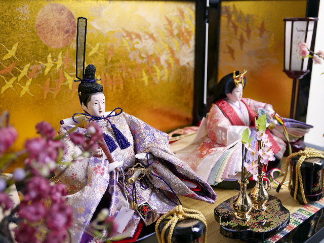 ほのかな赤と紫を組み合わせた優雅な光沢衣装の雛人形を鶴の金屏風と黒塗り台に並べる収納飾り